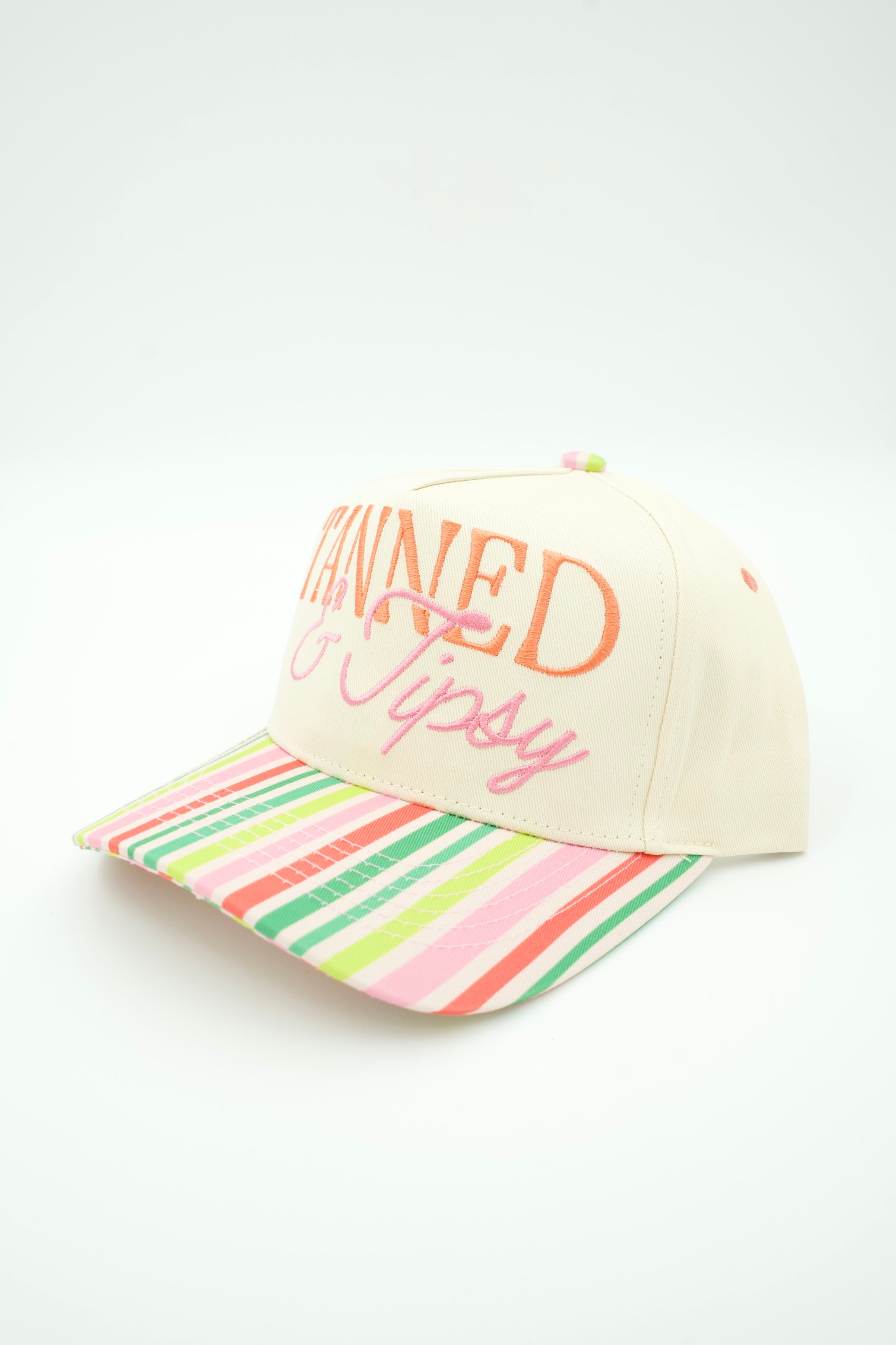 A cream-colored baseball cap with orange and pink embroidered text reading 'TANNED & Tipsy' on the front, featuring a vibrant multicolor striped brim.