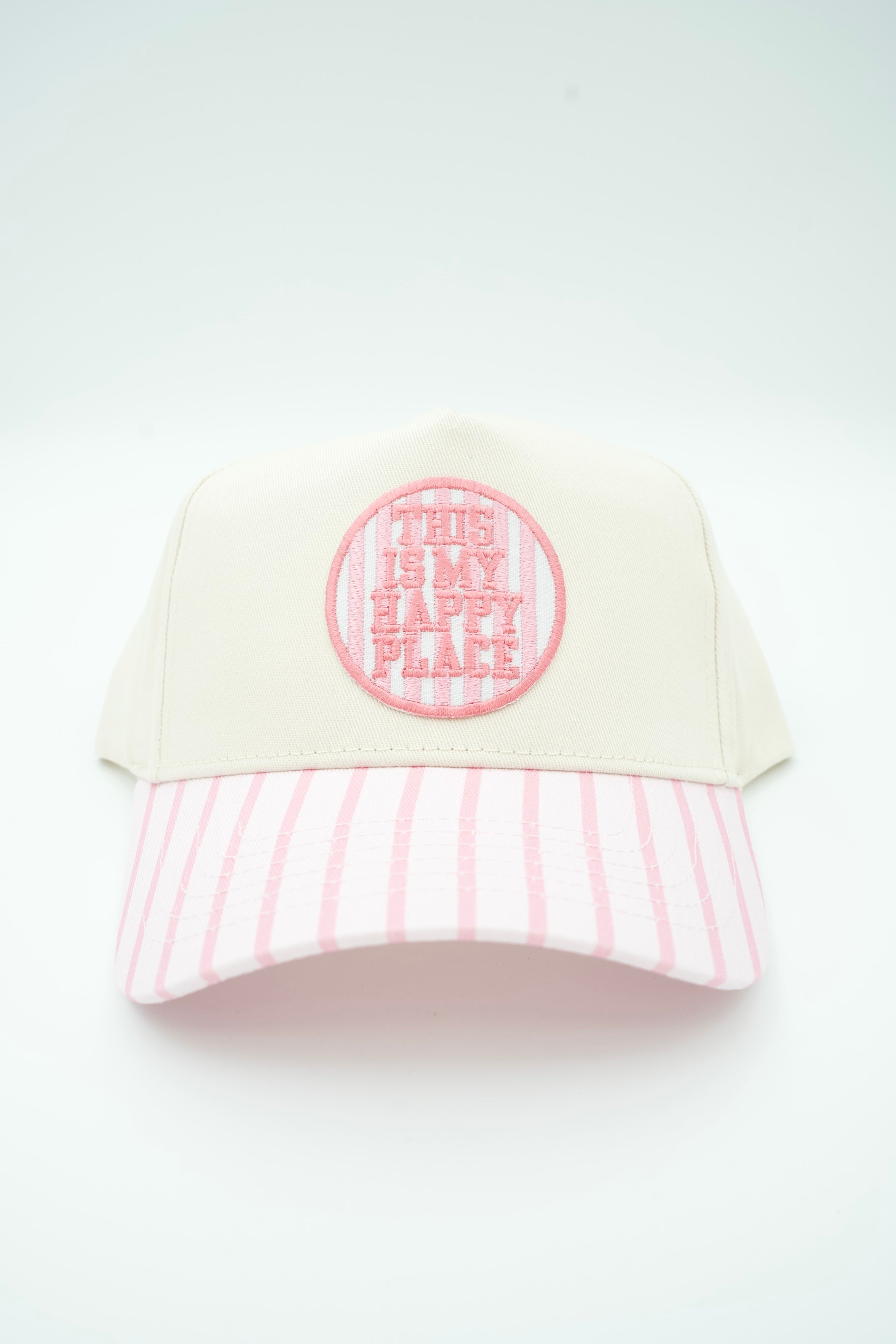 A baseball cap with an off-white crown, a pink and white striped visor, and a circular pink embroidered patch on the front that reads 'THIS IS MY HAPPY PLACE'.