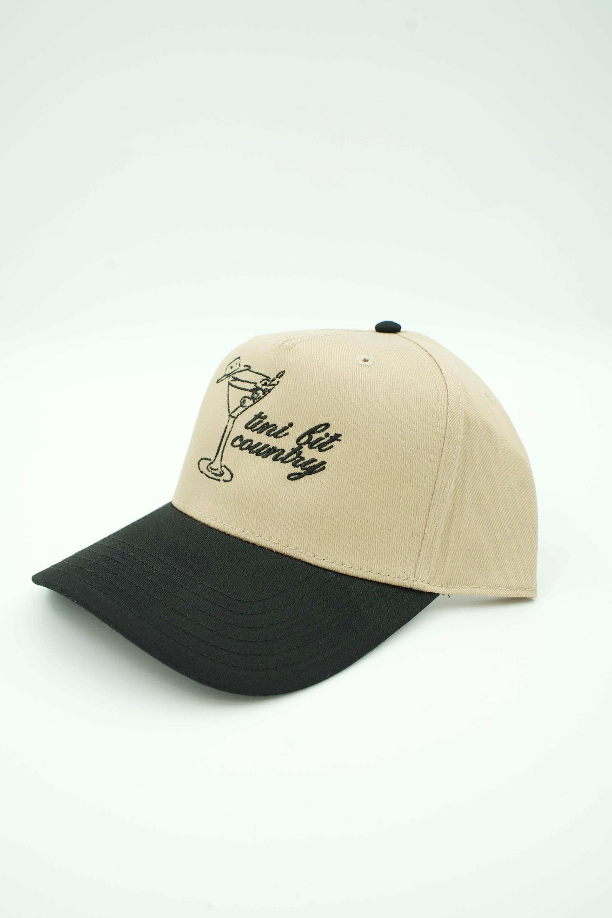 A two-tone baseball cap with a beige crown and a black curved brim, featuring a black embroidered graphic of a cocktail glass and the text 'mini bit country' on the front.