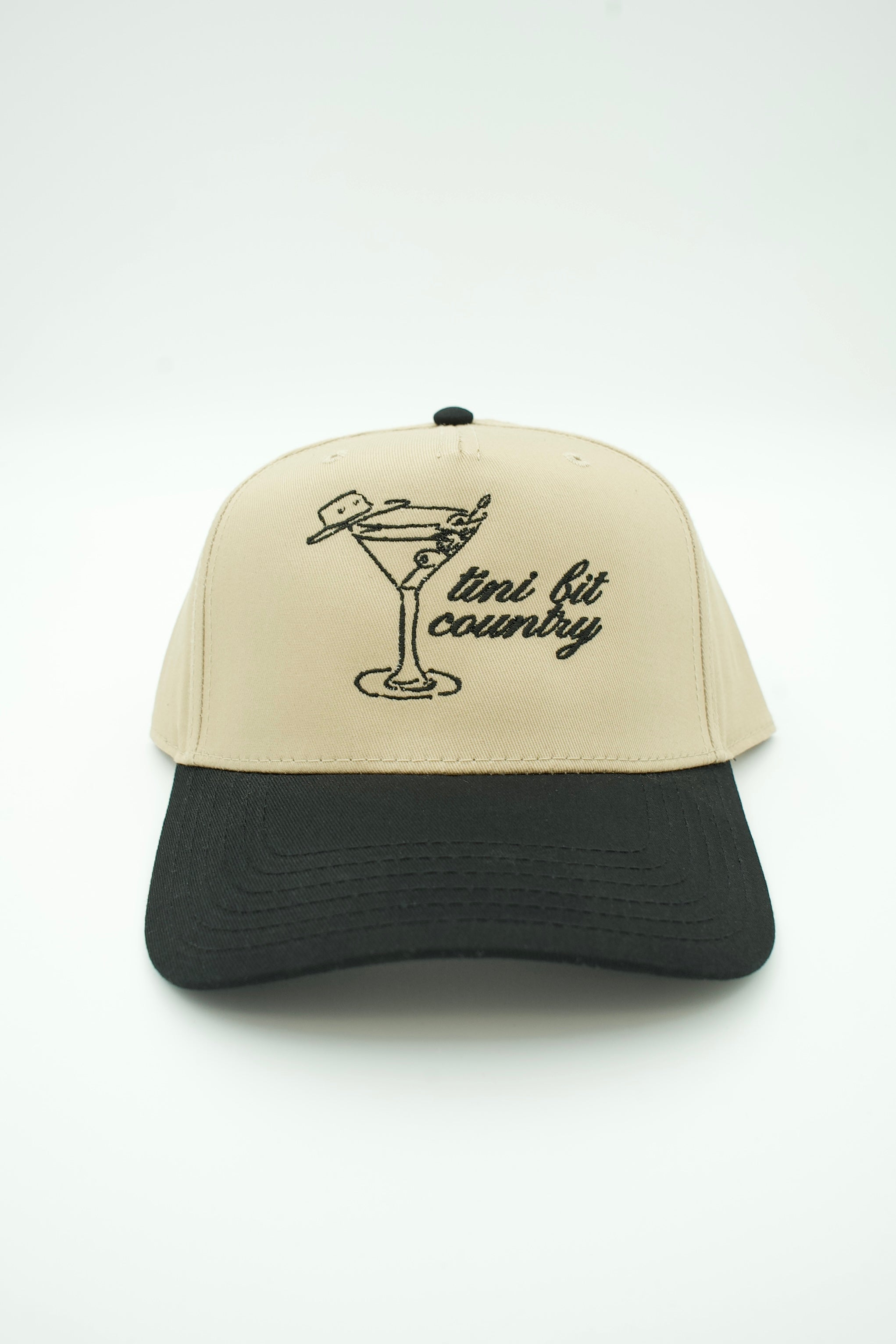 A two-tone baseball cap with a beige crown and a black curved brim, featuring a black embroidered graphic of a martini glass and the text 'mini bit country' on the front.