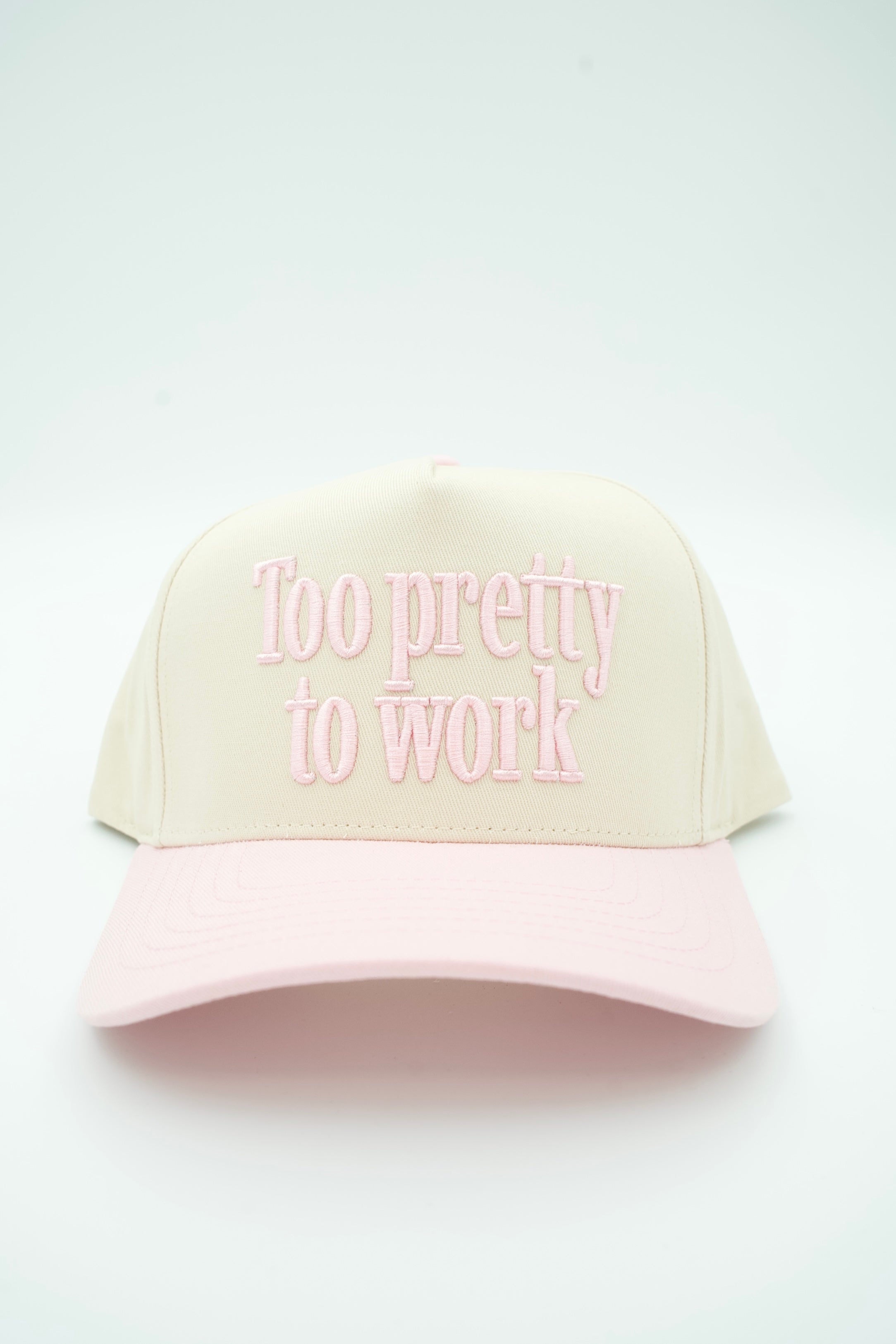 A cream-colored trucker cap with a light pink brim and pink embroidered text reading 'Too pretty to work' on the front, set against a plain white background.