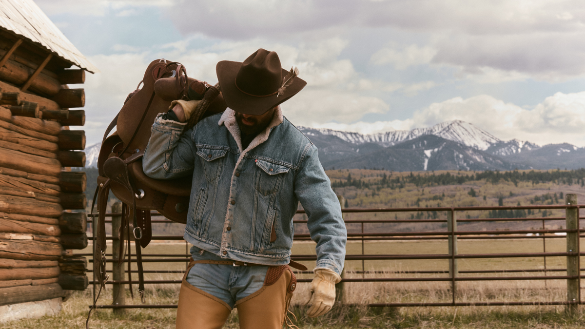 A person wearing a brown cowboy hat, a light blue denim jacket with a shearling lining, and tan leather chaps stands in front of a rustic log cabin with mountains in the background, carrying a large brown leather saddle on their back.