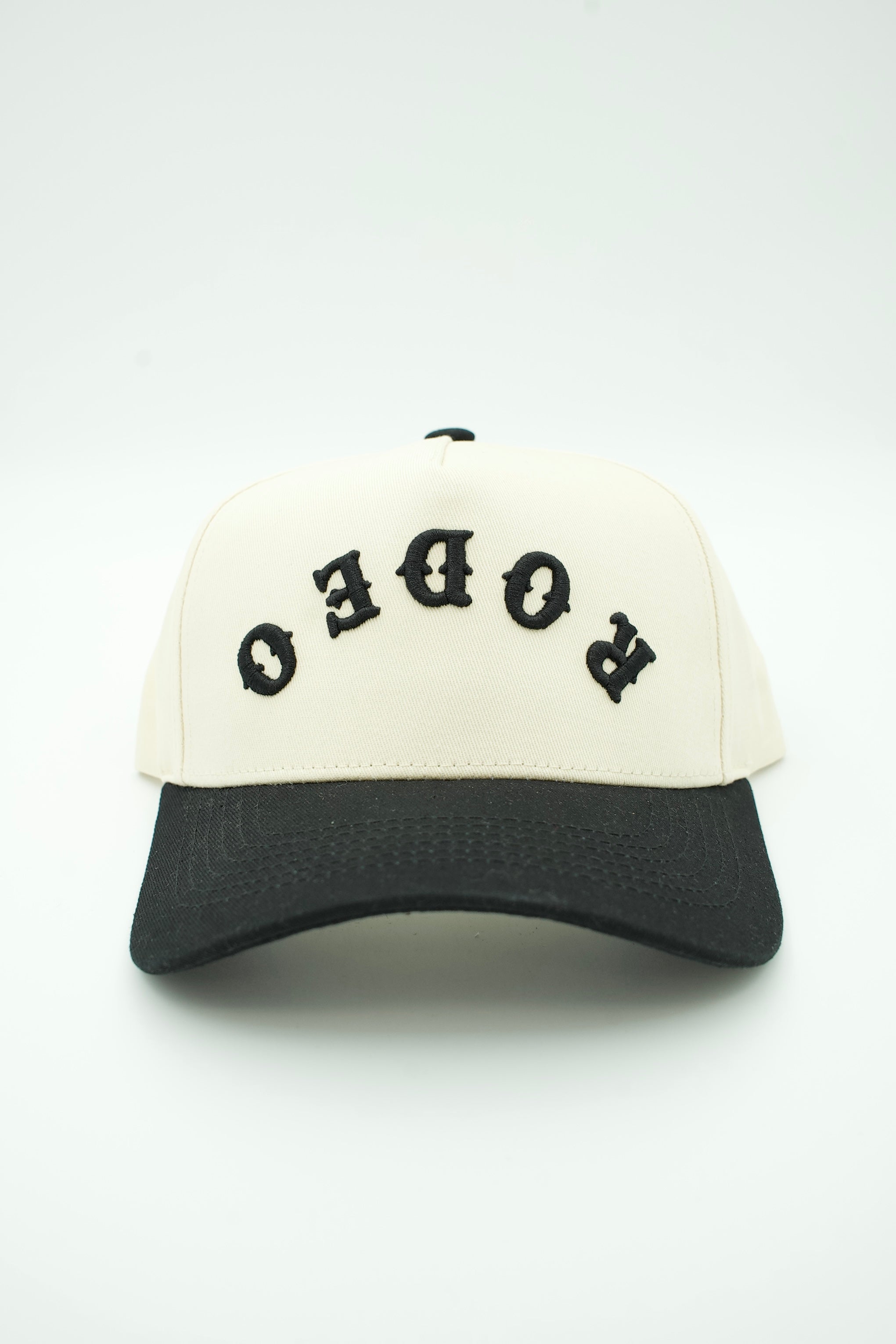 A two-tone baseball cap with an off-white crown and a black curved brim. The front of the cap features the word 'RODEO' embroidered in black gothic-style letters, which are upside down relative to the front view.