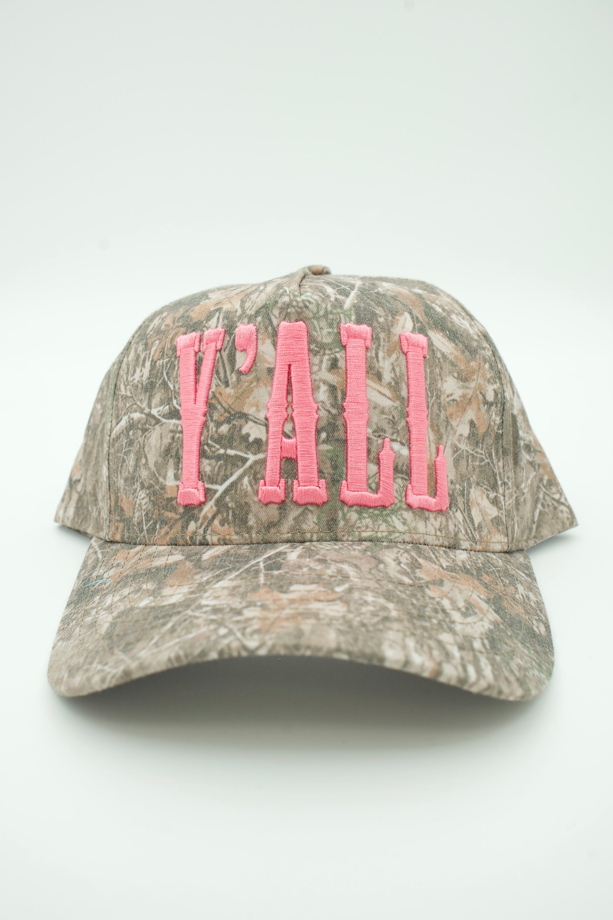 A baseball cap featuring a brown and gray woodland camouflage pattern with pink embroidered text reading 'Y'ALL' on the front, set against a plain light gray background.