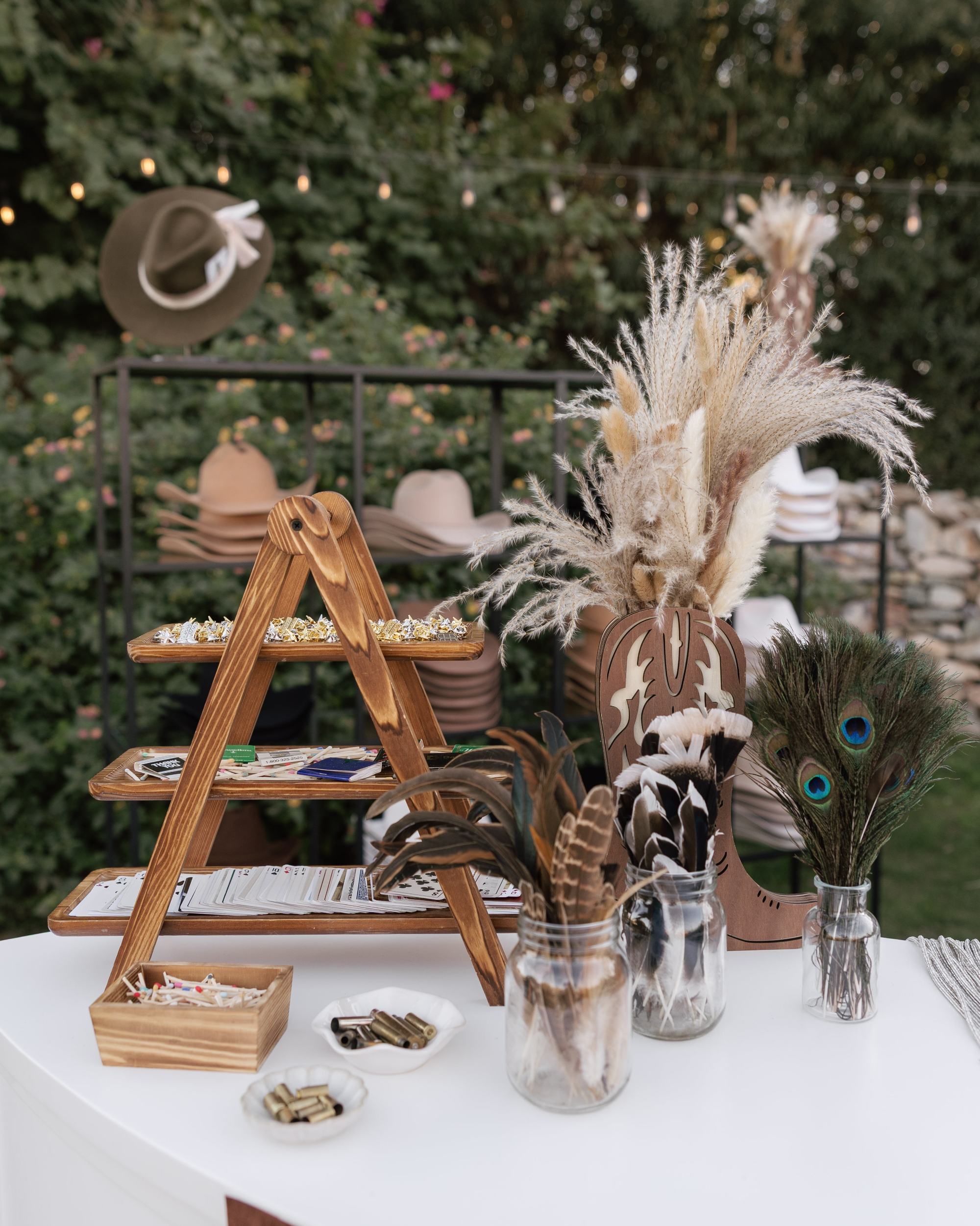 Decorative table setup with a wooden tiered stand, jars with feathers, and a hat in an outdoor setting.